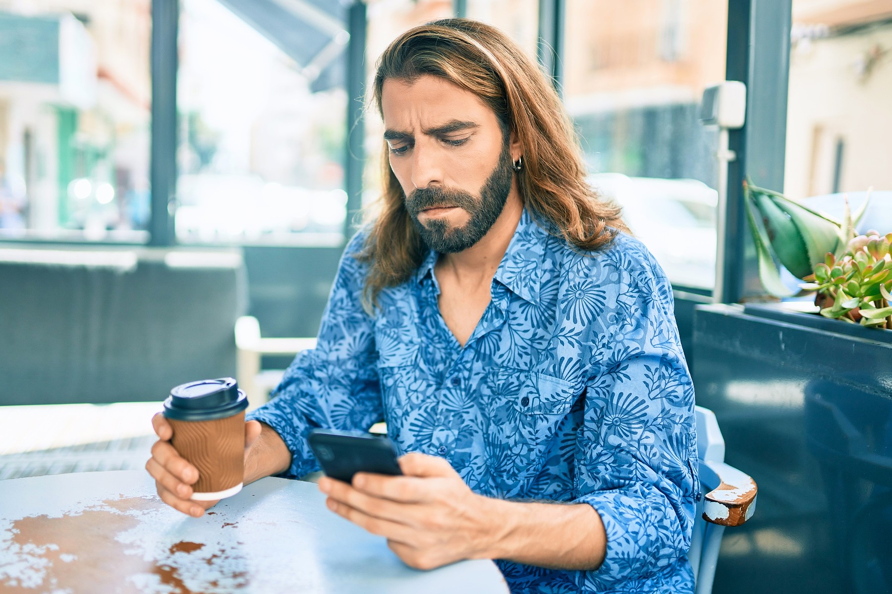 A man with long hair and a beard, wearing a blue patterned shirt, sits at a table in a cafe holding a coffee cup and looking at his smartphone with a serious expression.