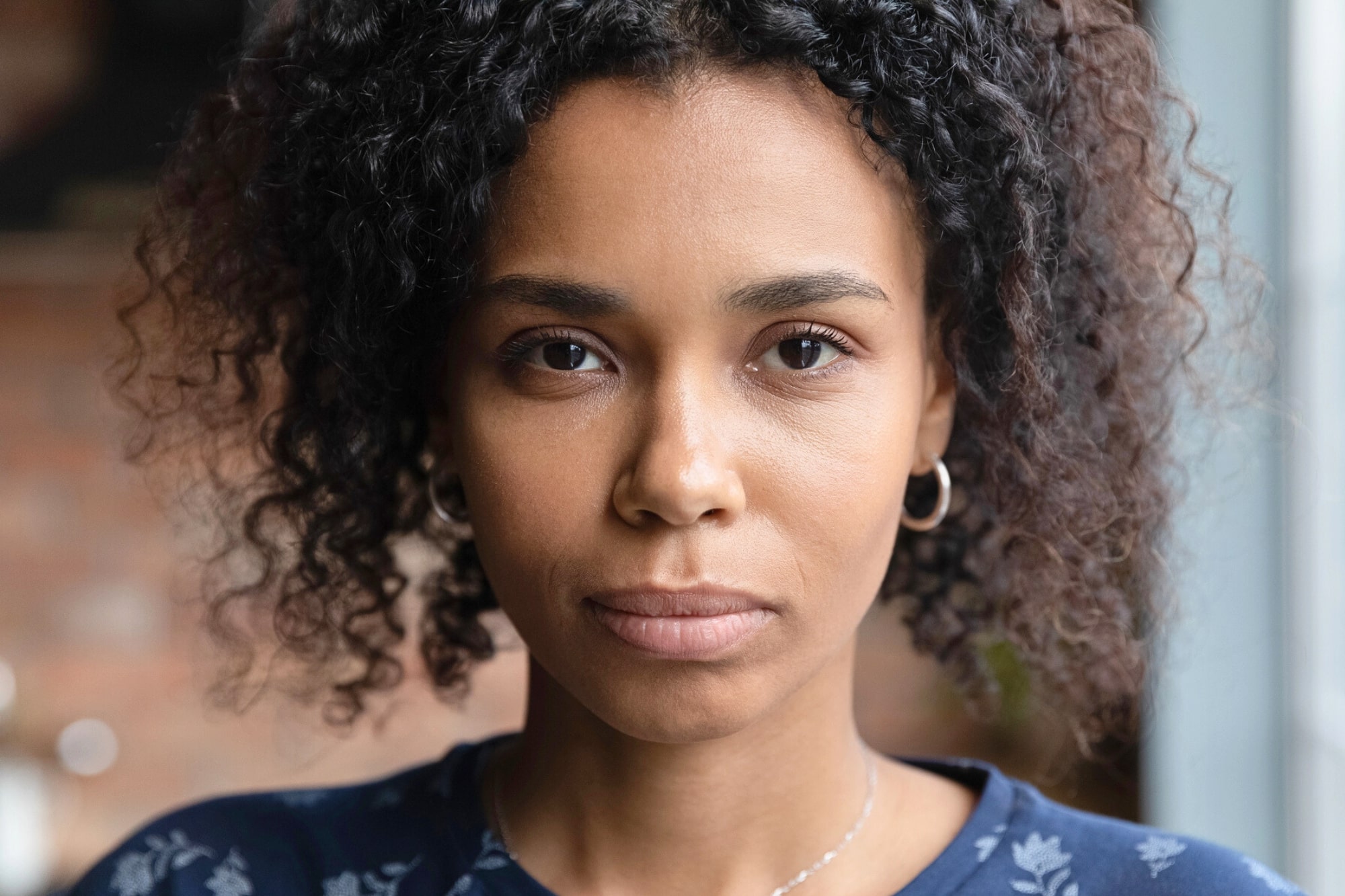 A woman with curly dark hair and hoop earrings looks directly at the camera with a neutral expression, wearing a blue top with a subtle floral pattern. The background is softly blurred.