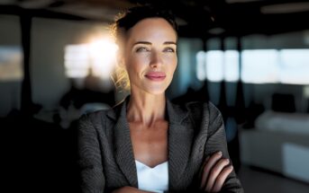 Confident woman in a grey blazer stands with arms crossed, smiling slightly. Sunlight streams through windows behind her in a modern office setting, creating a warm and professional atmosphere.