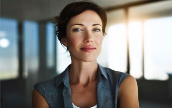 A confident woman with short brown hair and blue eyes stands indoors, smiling slightly, with sunlight streaming through large windows in the background.