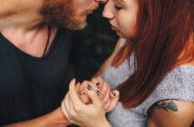 A close-up of a couple embracing tenderly, holding hands and touching foreheads. The woman has red hair and a tattoo on her arm; the man has a short beard. Their eyes are closed, conveying intimacy and affection.