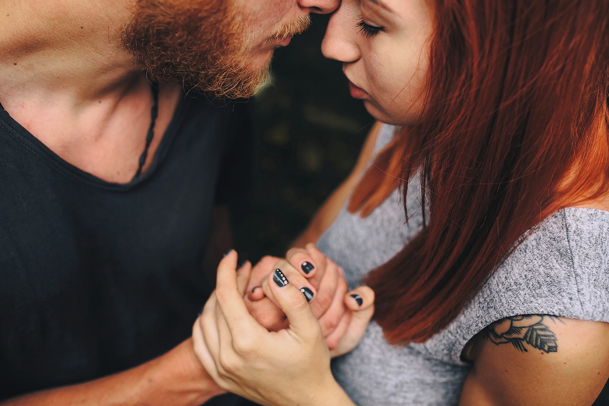 A close-up of a couple embracing tenderly, holding hands and touching foreheads. The woman has red hair and a tattoo on her arm; the man has a short beard. Their eyes are closed, conveying intimacy and affection.