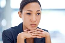 A woman in a dark blazer rests her chin on her hands, looking thoughtful and serious. The background is softly blurred, drawing focus to her contemplative expression.