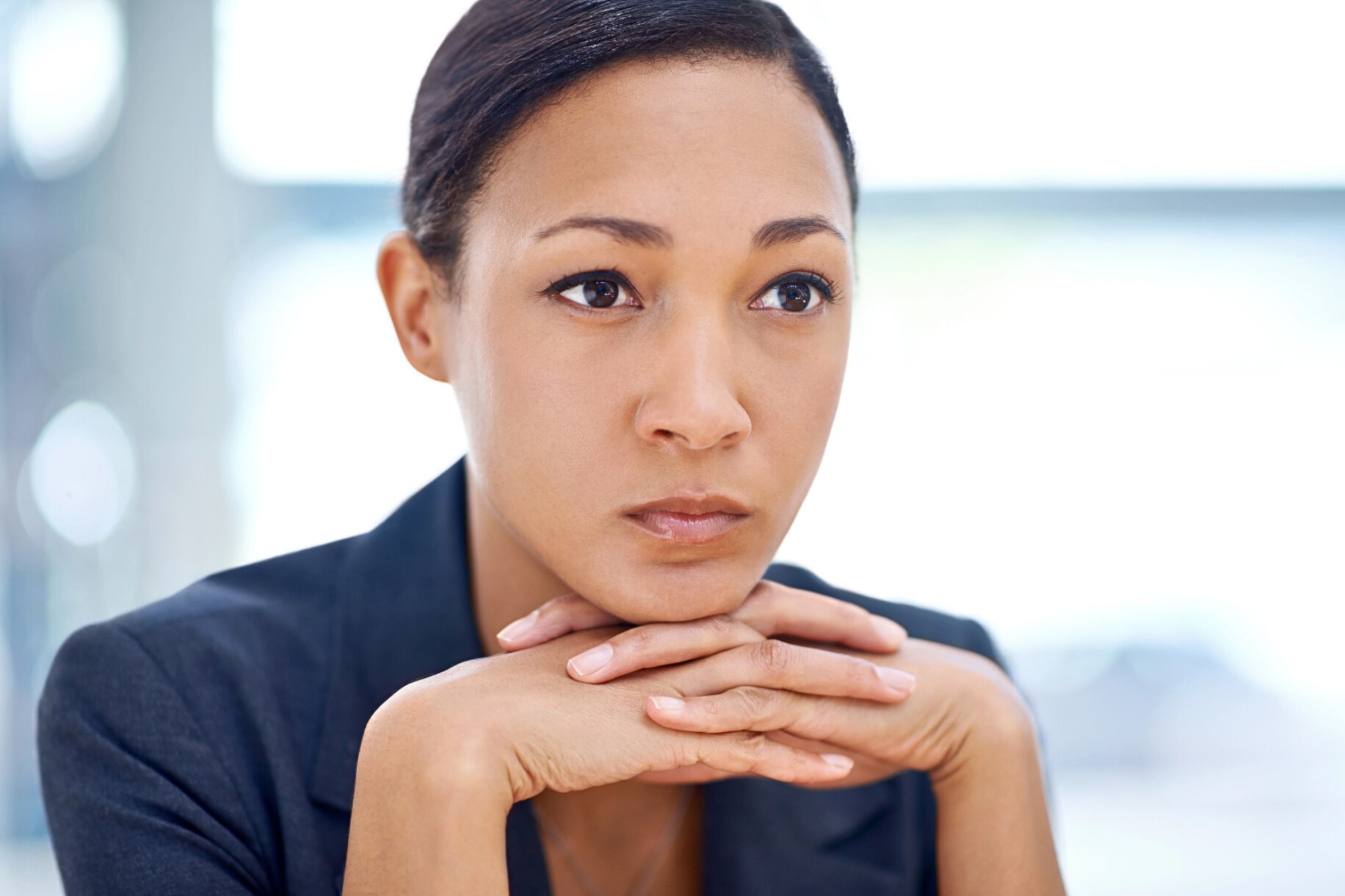 A woman in a dark blazer rests her chin on her hands, looking thoughtful and serious. The background is softly blurred, drawing focus to her contemplative expression.