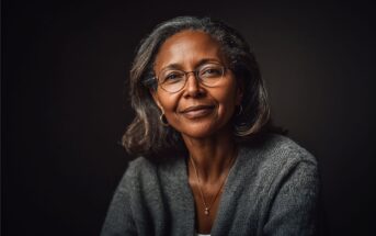 A middle-aged woman with gray hair and glasses smiles gently, wearing a gray sweater and a necklace, posed against a dark background.