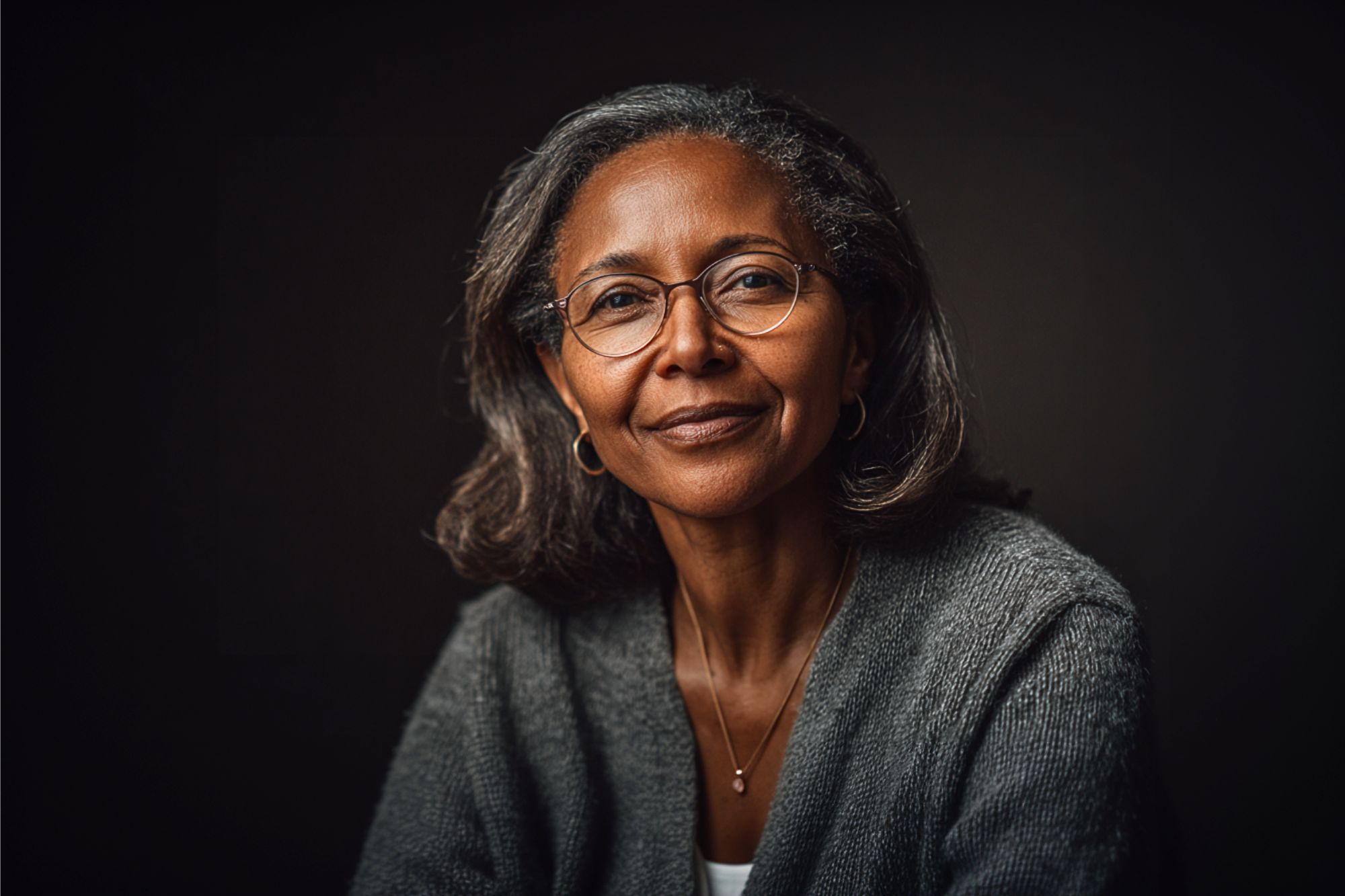 A middle-aged woman with gray hair and glasses smiles gently, wearing a gray sweater and a necklace, posed against a dark background.