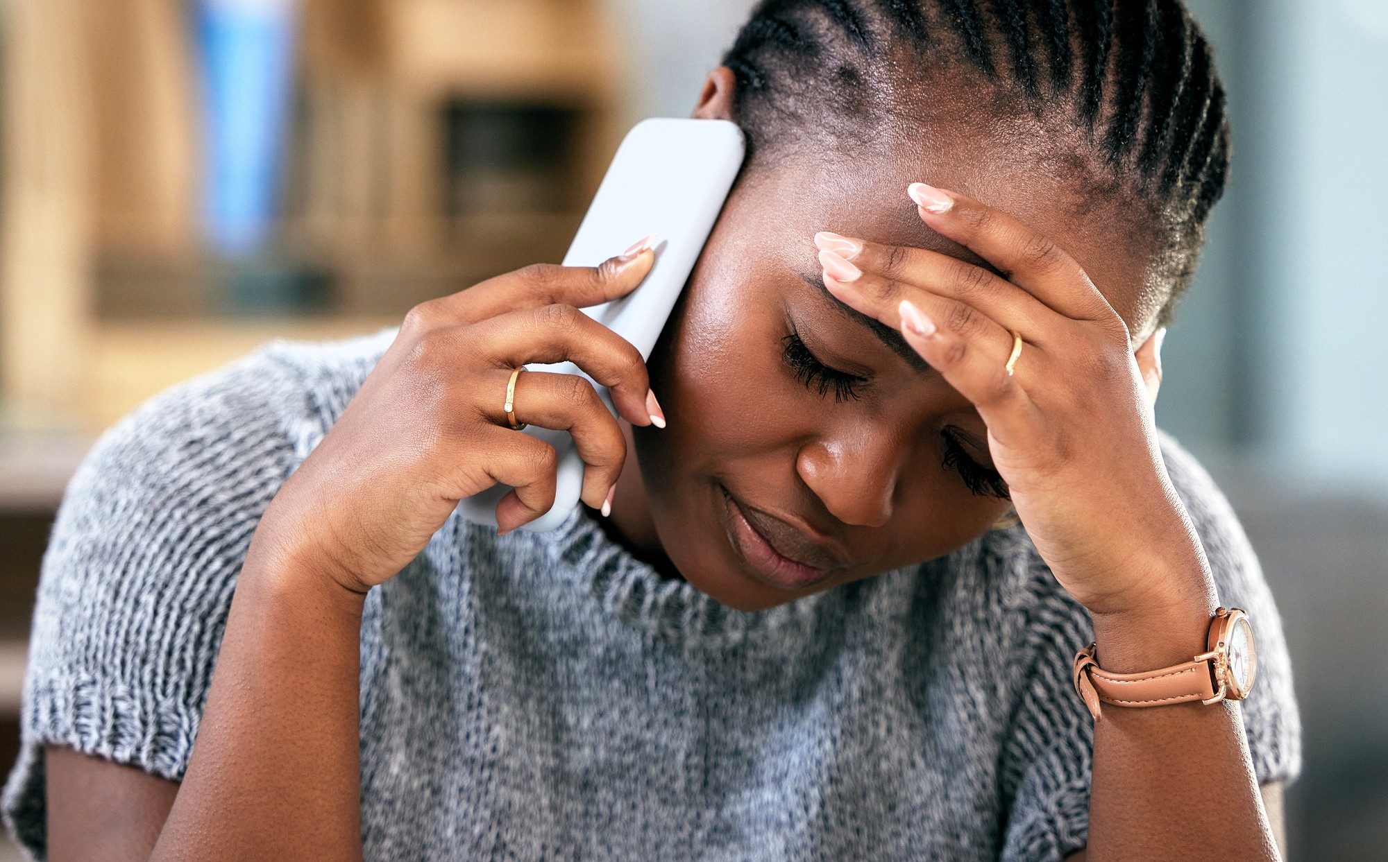 A woman with braided hair looks stressed while talking on the phone, holding her forehead with one hand and her phone with the other. She wears a gray sweater and a watch, and sits indoors.