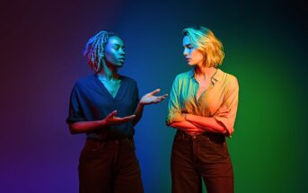 Two women stand facing each other under dramatic, colorful lighting. One gestures with her hands while speaking; the other stands with arms crossed, listening. The background blends blue, red, and green hues.