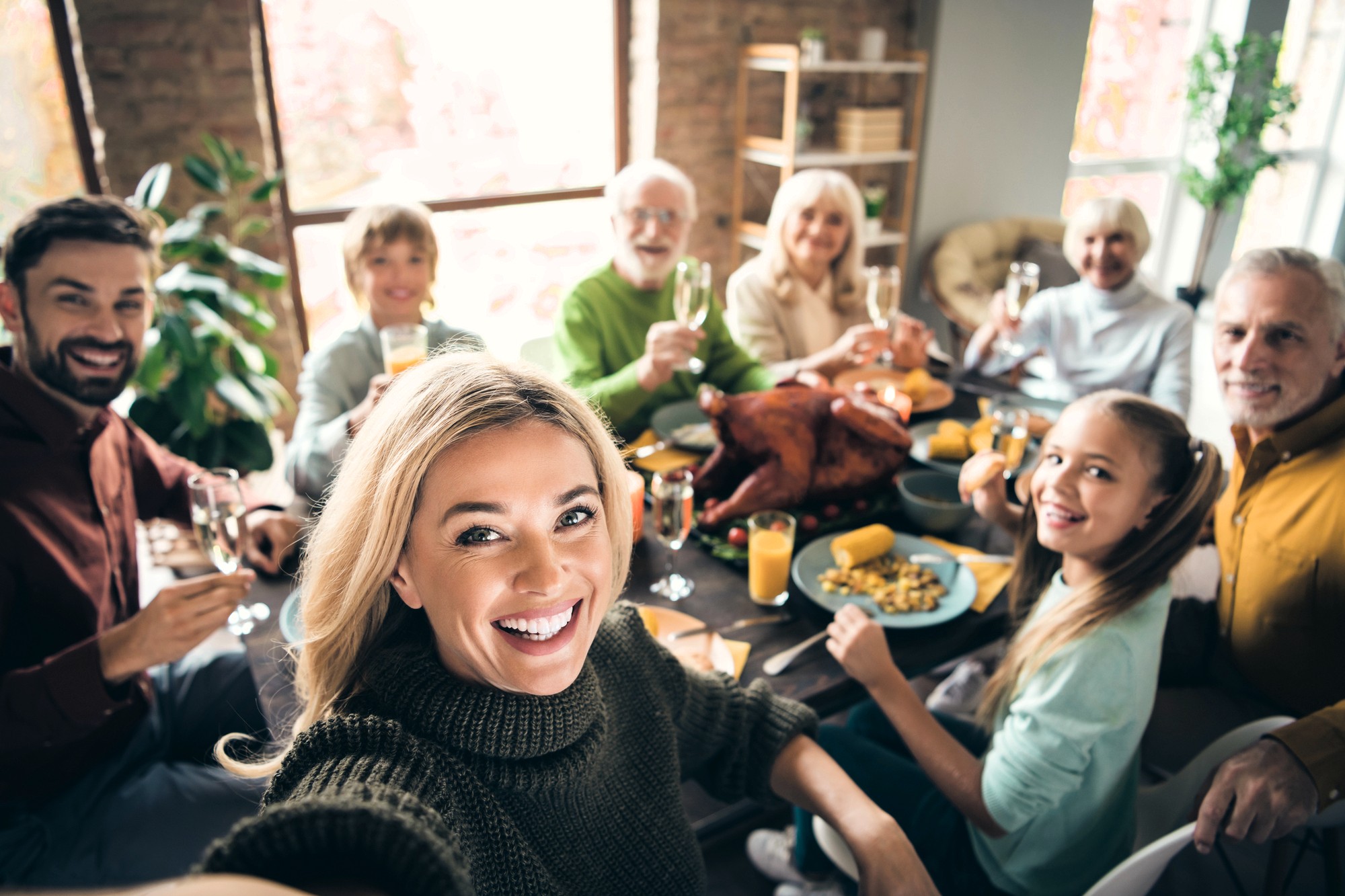 A group of people of various ages sit around a dinner table with a roast turkey, smiling and raising glasses, while a woman in front takes a selfie. The scene appears cheerful and festive.