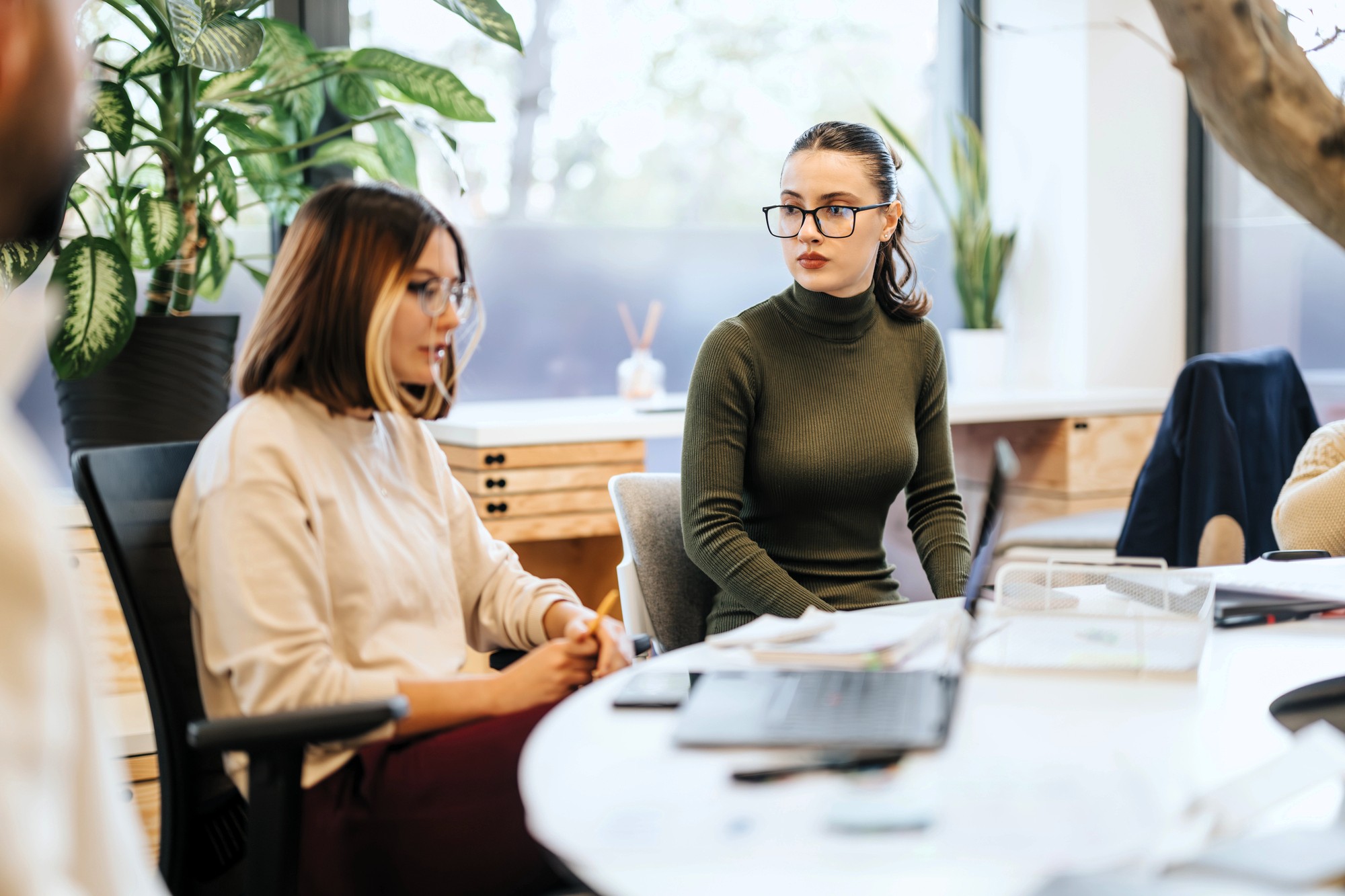 Two women sitting at a desk in a modern office, having a discussion. One is wearing a white shirt, the other a green turtleneck. Papers, a laptop, and office supplies are on the table, with plants and large windows in the background.