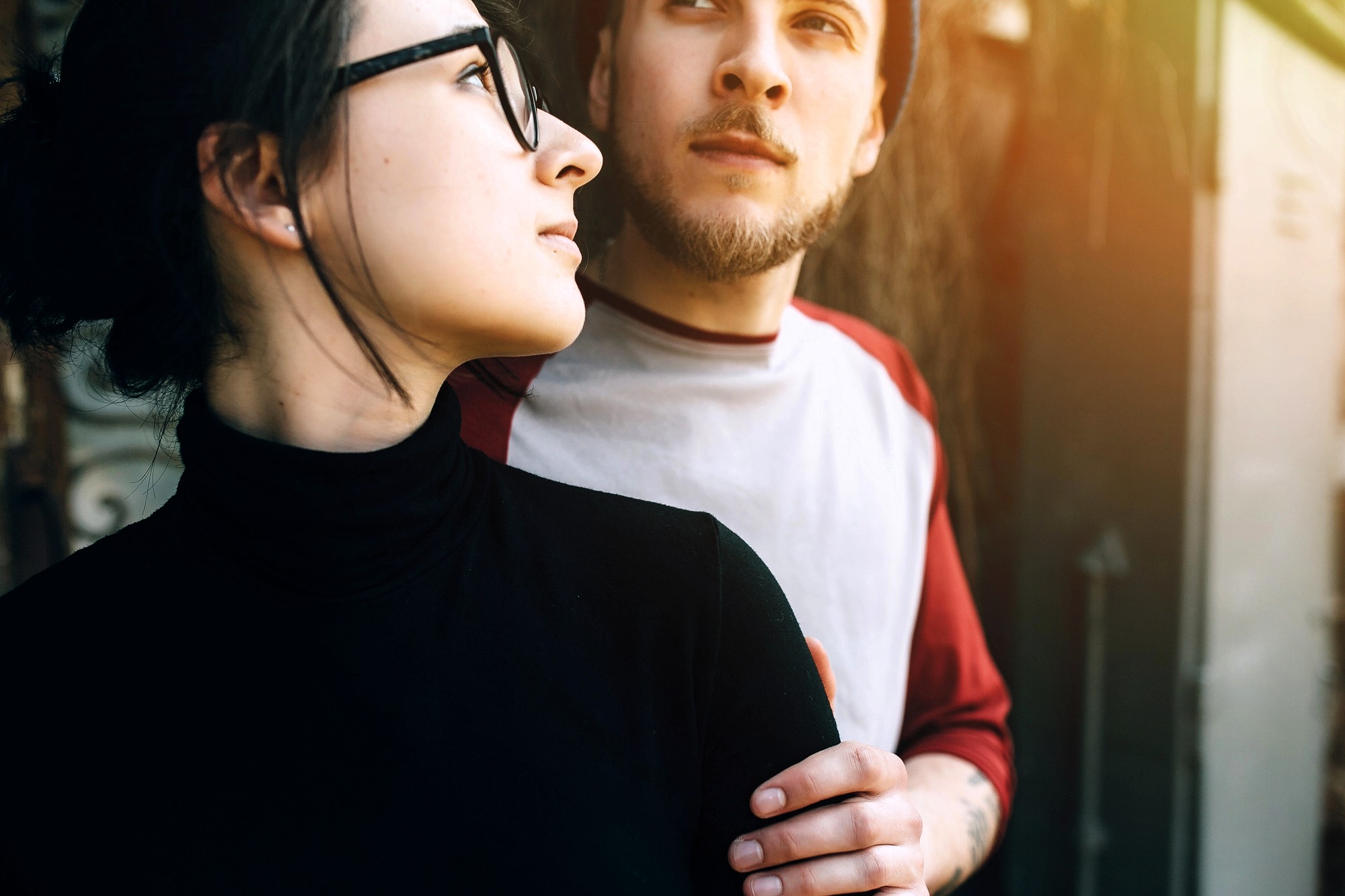 A woman in glasses and a black turtleneck stands closely with a man in a white and red shirt, gently holding his arm. Both look thoughtfully into the distance, illuminated by soft sunlight.