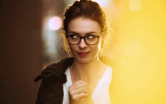 A woman with brown hair and glasses stands in soft lighting, looking to her left with a playful, mischievous expression, hands clasped under her chin. A warm yellow light fills the right side of the image.