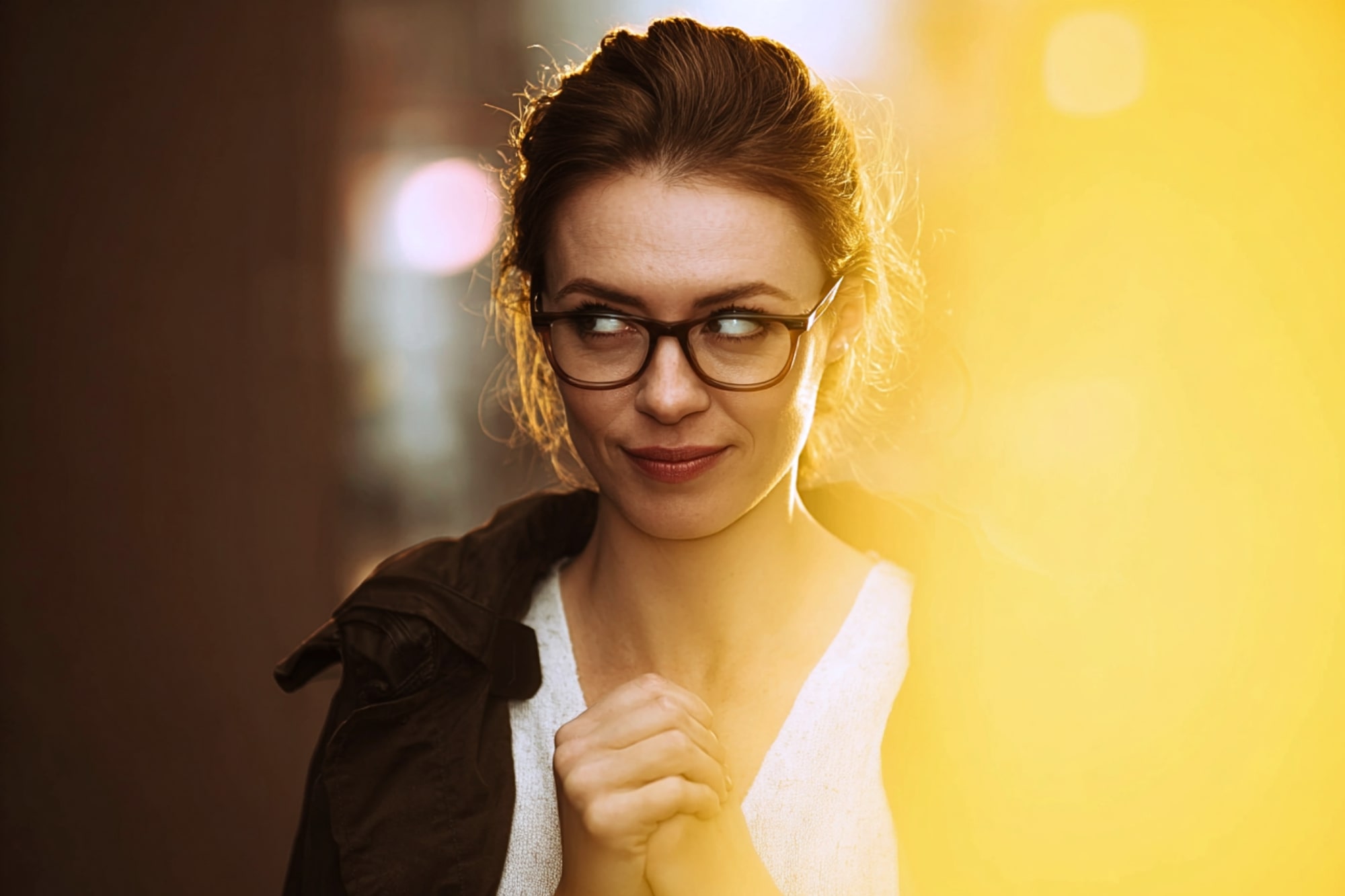 A woman with brown hair and glasses stands in soft lighting, looking to her left with a playful, mischievous expression, hands clasped under her chin. A warm yellow light fills the right side of the image.