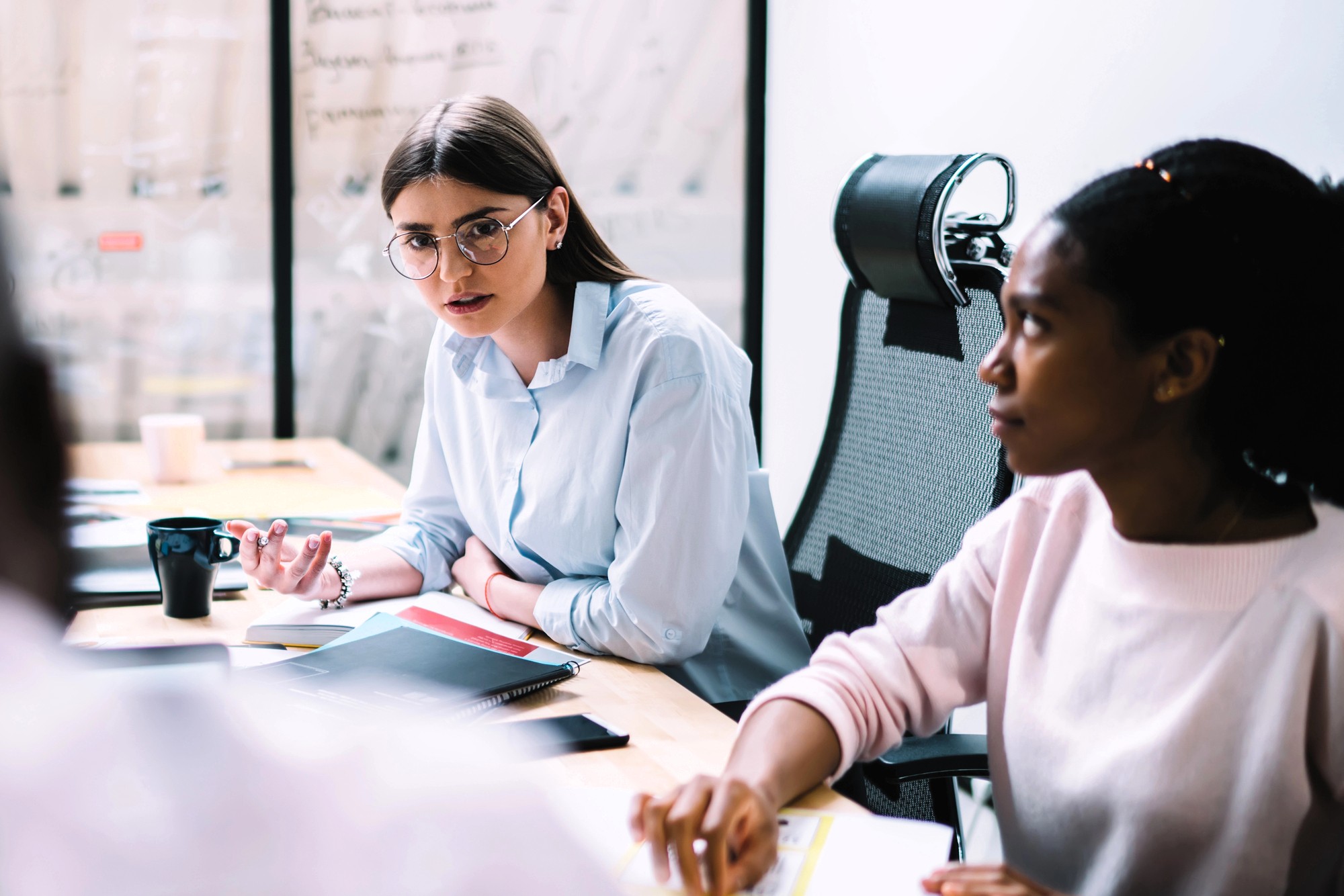 Two women sit at a conference table in a modern office, engaged in discussion. One woman with glasses speaks, while the other listens attentively. Notebooks, documents, and a coffee mug are on the table.