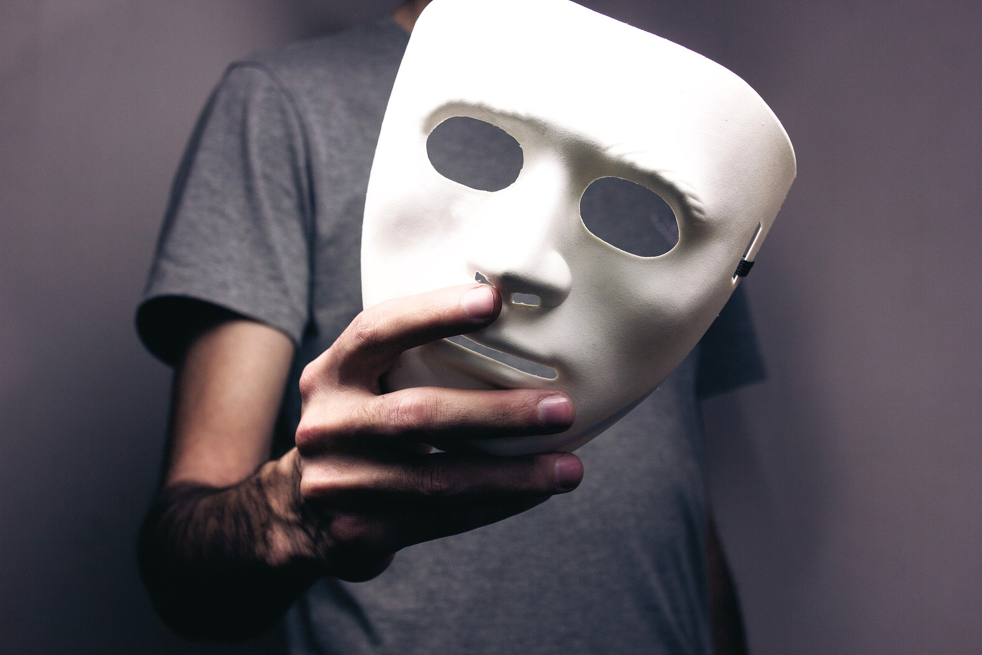 A person wearing a gray shirt holds a blank white mask in front of their chest, with only their hand and part of their upper body visible.