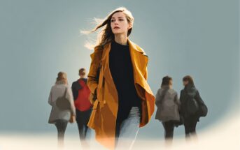 A woman in a yellow coat walks confidently toward the viewer, with wind blowing her hair, while four blurred figures walk in the opposite direction in the background against a soft, grayish-blue backdrop.