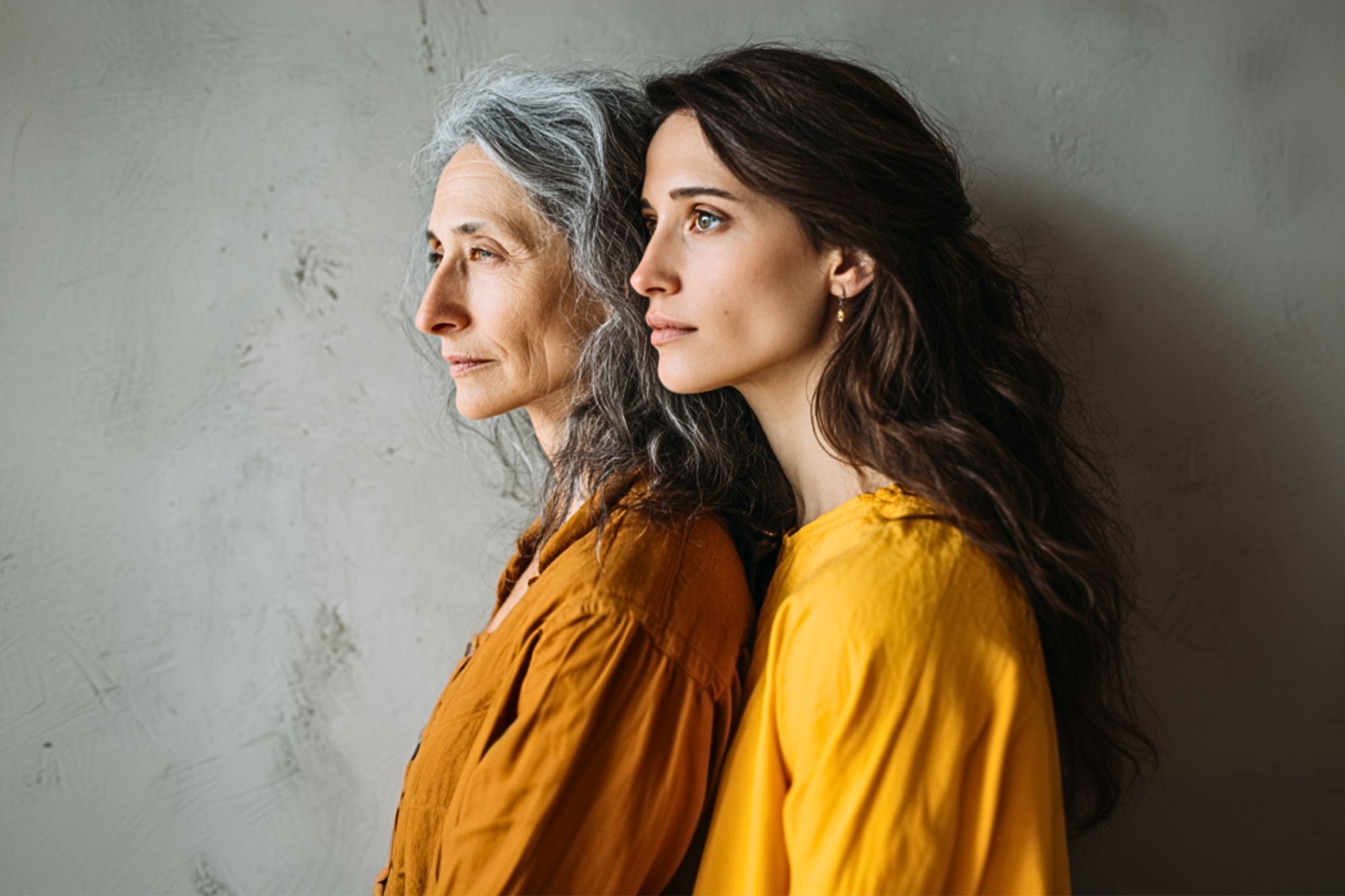 Two women, one older with gray hair and one younger with long brown hair, stand side by side in profile, both wearing mustard yellow tops against a neutral textured background.