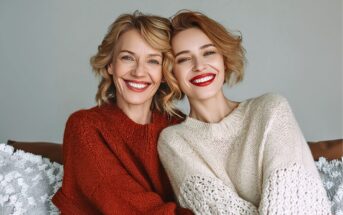 Two women with short, wavy blonde hair are sitting close together, smiling warmly at the camera. One wears a red sweater and the other a cream one. They appear happy and relaxed against a neutral background.