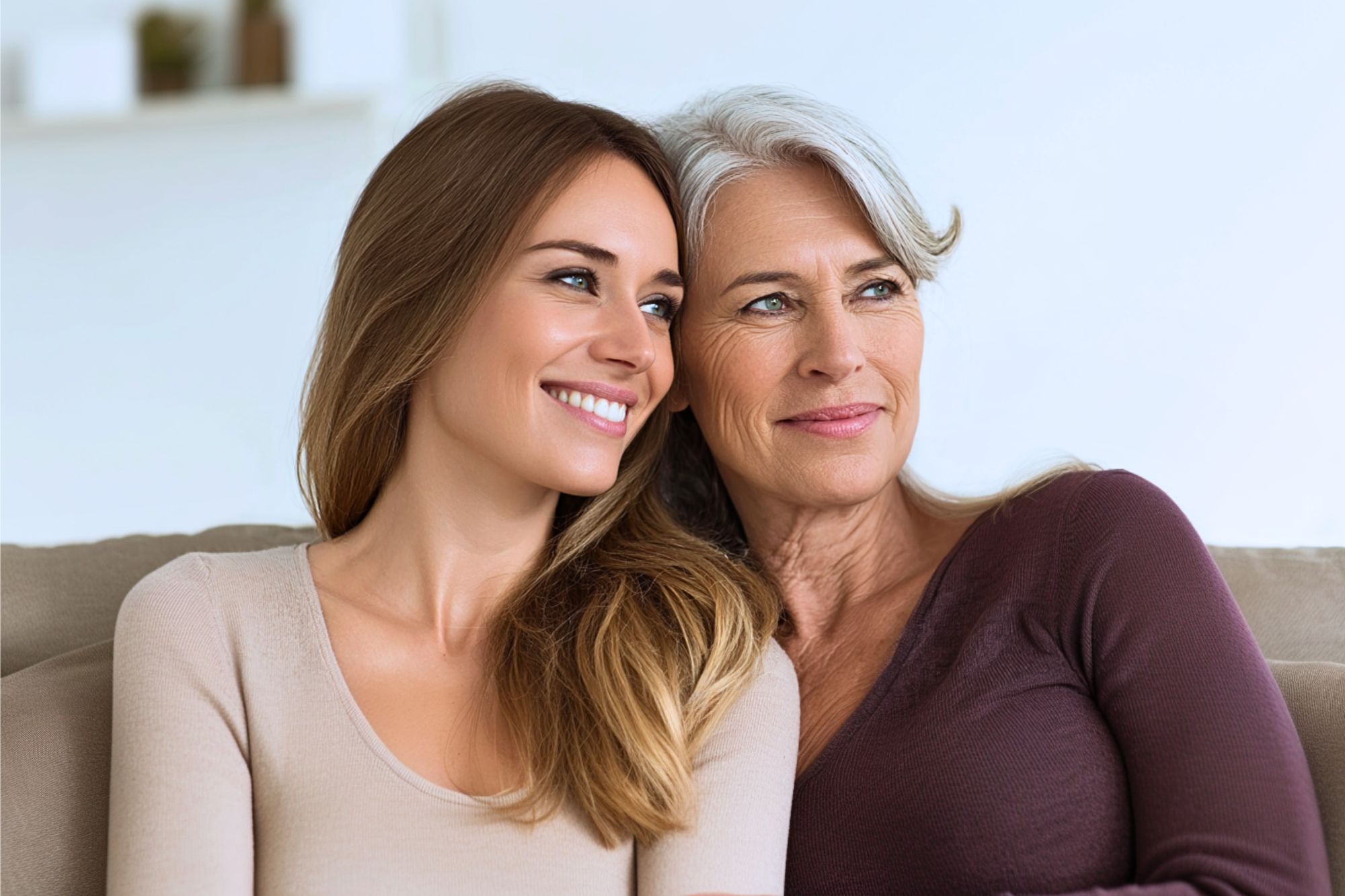 Two women, one younger with long brown hair and one older with gray hair, are sitting close together on a couch, smiling and looking off to the side.
