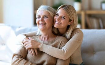 A young woman smiling while embracing her smiling older mother from behind on a sofa.
