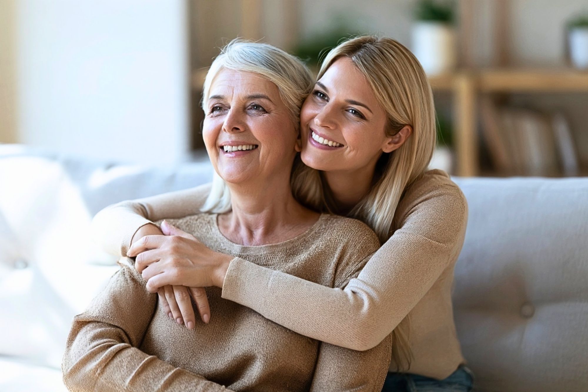 A young woman smiling while embracing her smiling older mother from behind on a sofa.
