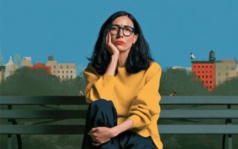 A woman with dark hair and glasses wears a yellow sweater and sits on a park bench in front of a city skyline.