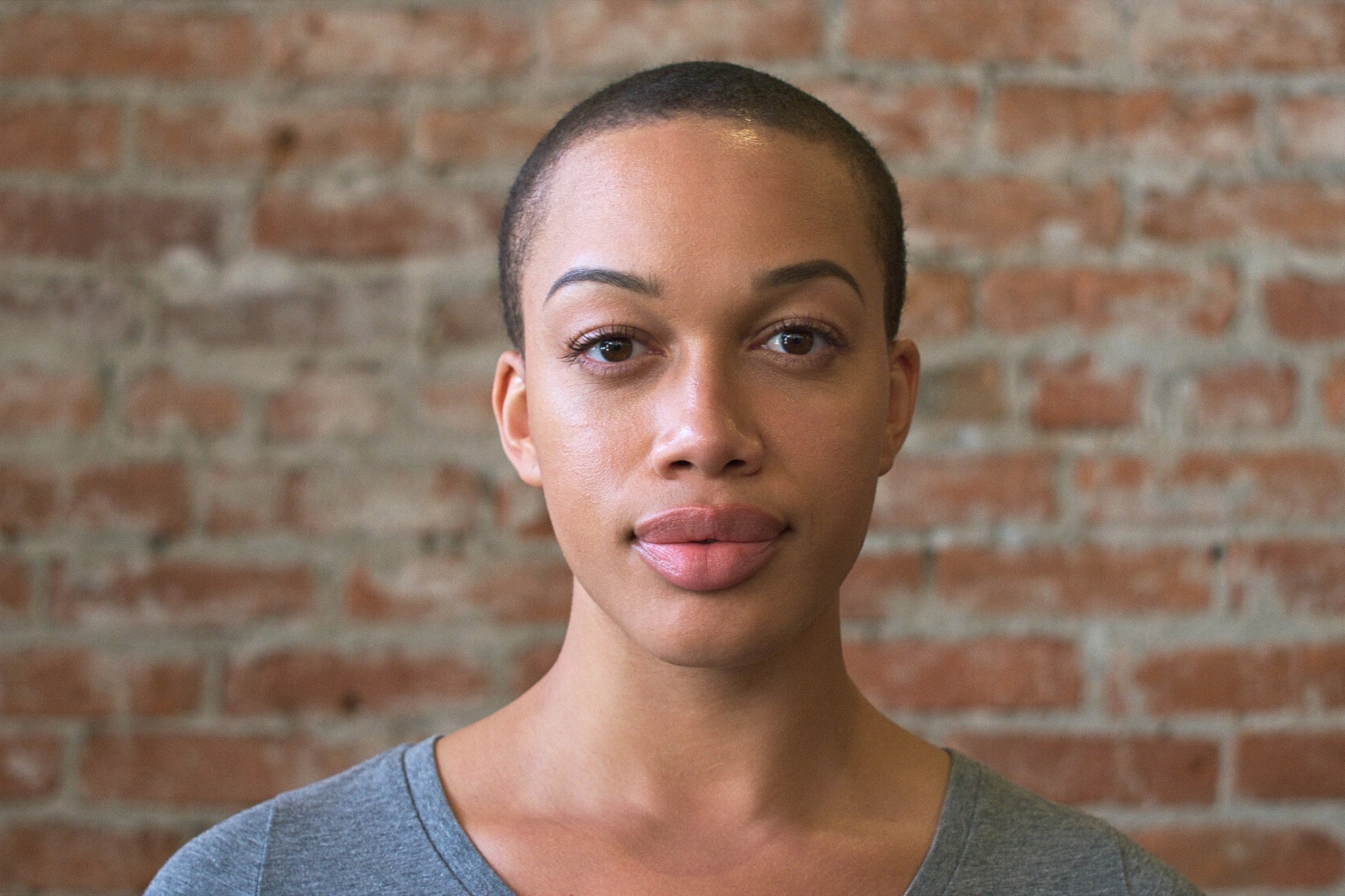 A person with short hair and a gray shirt stands in front of a brick wall, looking directly at the camera with a neutral expression.