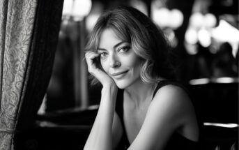 A woman with wavy hair sits by a window, resting her head on her hand and smiling softly. The image is in black and white, with a blurred background and gentle light illuminating her face.