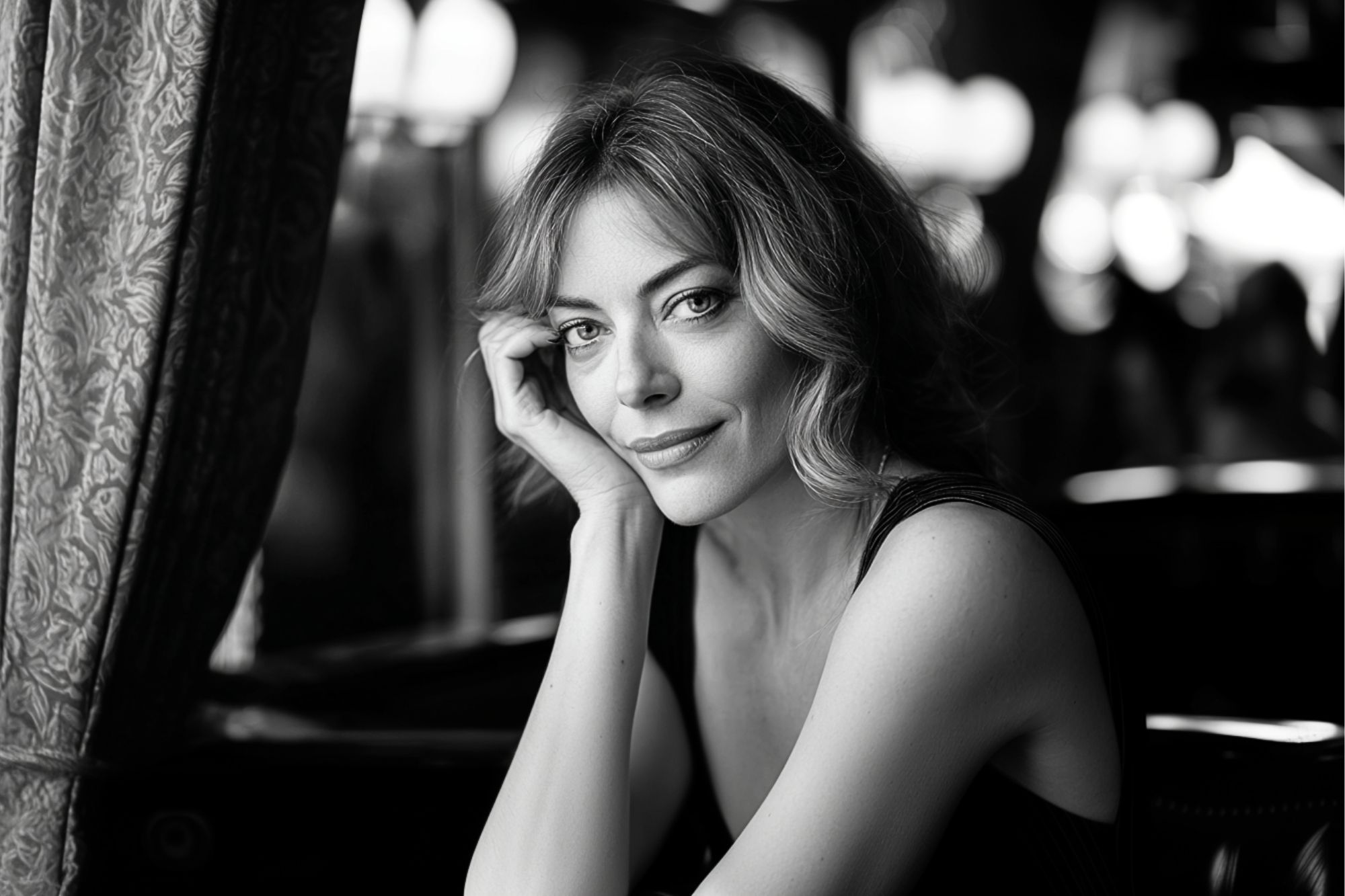 A woman with wavy hair sits by a window, resting her head on her hand and smiling softly. The image is in black and white, with a blurred background and gentle light illuminating her face.