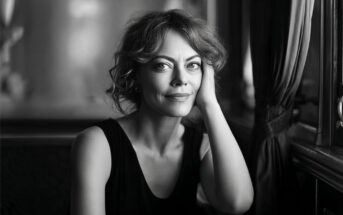 Black and white portrait of a woman with curly hair resting her chin on her hand, looking toward the camera.