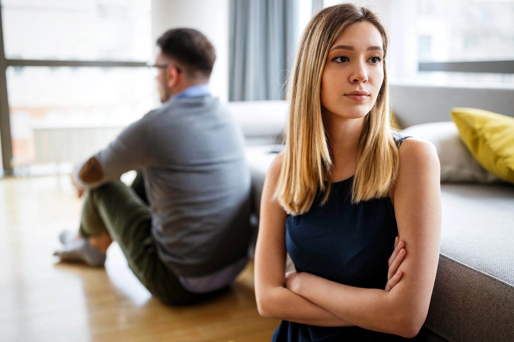 A young woman with crossed arms looks away, appearing upset, while a man sits on the floor with his back to her. They are indoors in a bright living room, suggesting tension or disagreement.