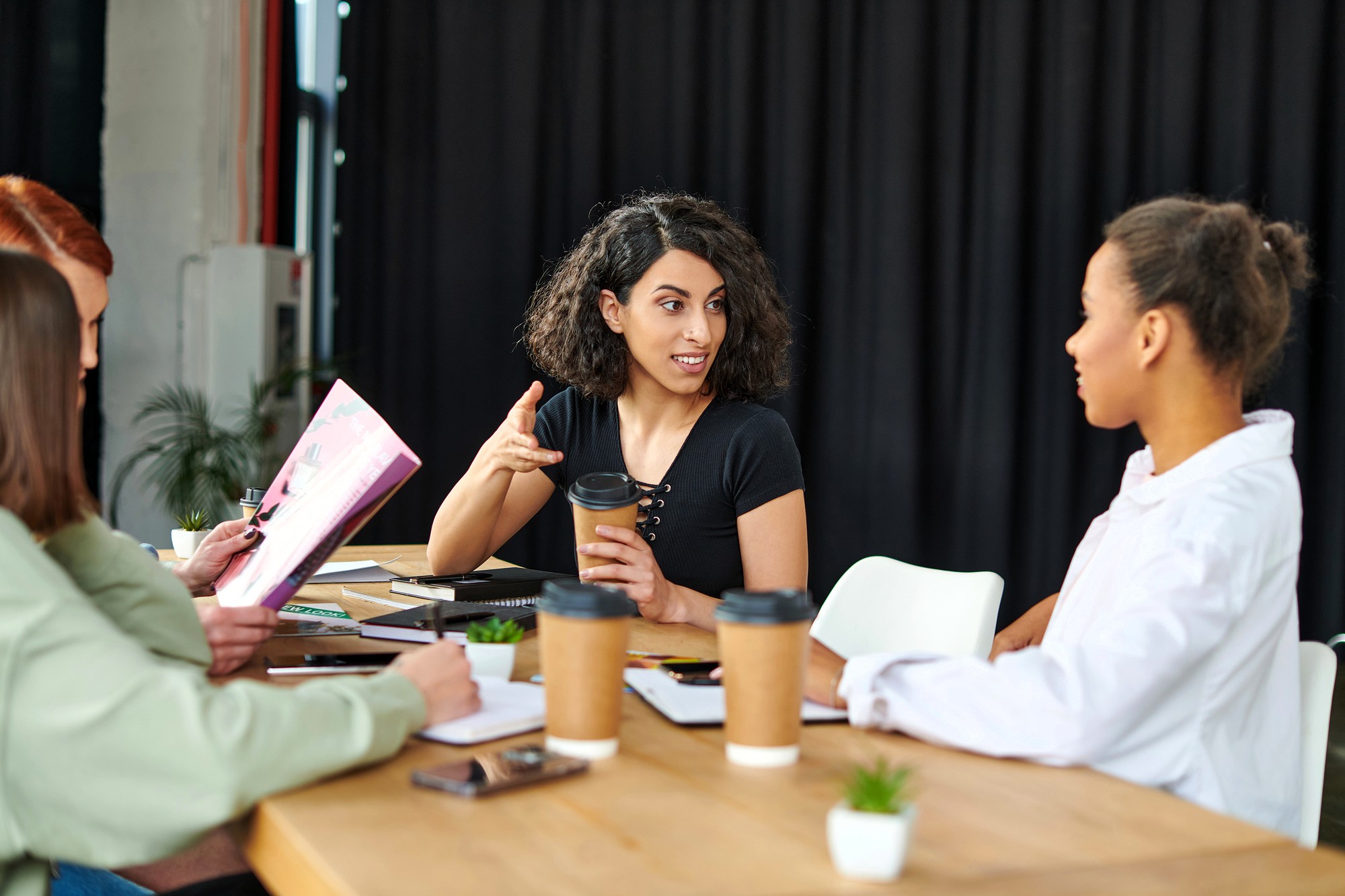 Four women sit at a table with coffee cups, notebooks, and a small plant, having a discussion. One woman gestures while speaking, and the others listen attentively. A dark curtain is in the background.