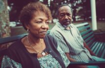 An older woman and man sit on a green park bench. The woman looks thoughtful or concerned, while the man gazes toward her. Trees and a grassy area are visible in the background.