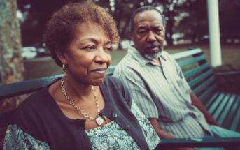 An older woman and man sit on a green park bench. The woman looks thoughtful or concerned, while the man gazes toward her. Trees and a grassy area are visible in the background.