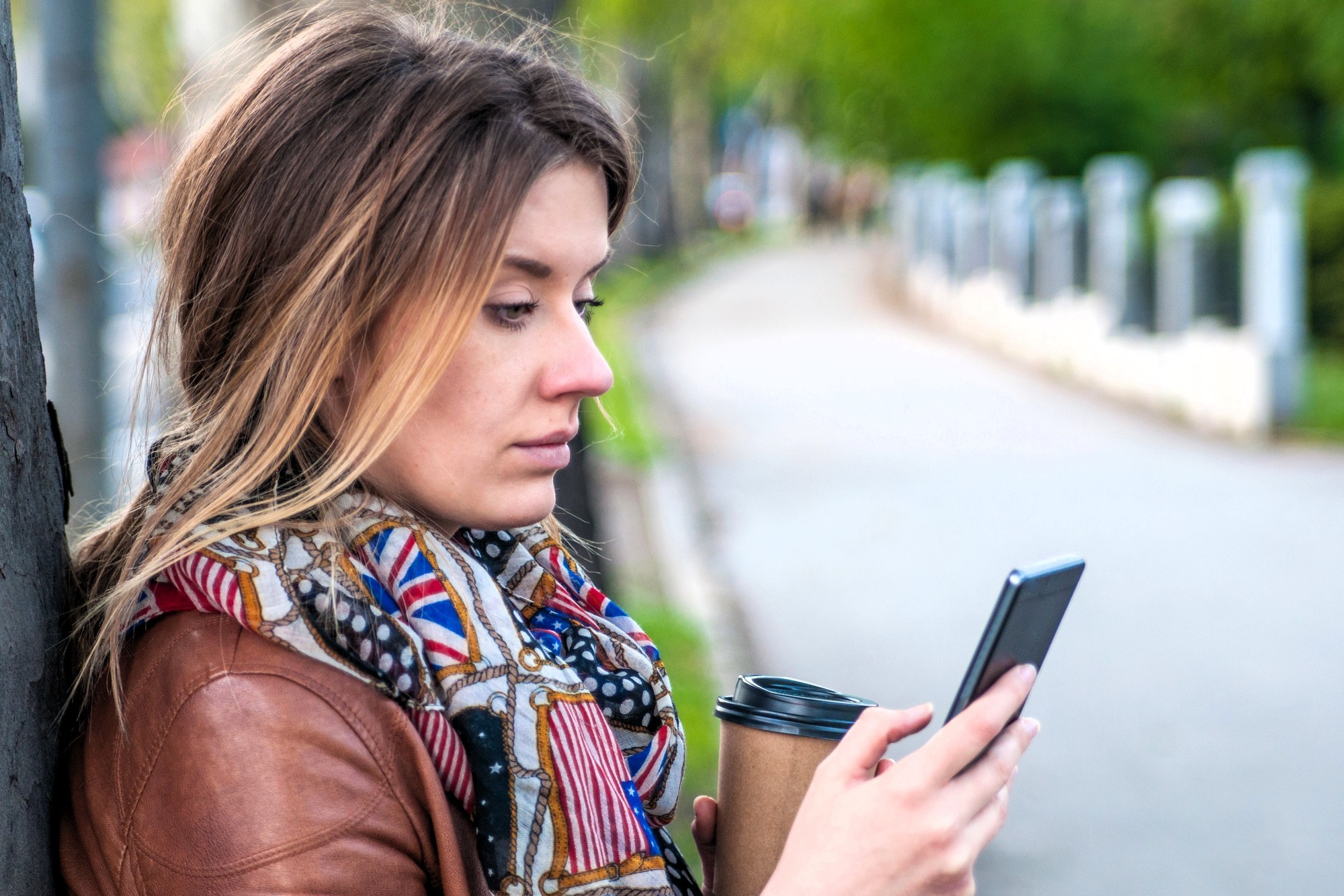 A woman with long hair, wearing a patterned scarf and brown jacket, stands outdoors holding a coffee cup and looking at her smartphone. Trees and a pathway are visible in the blurred background.