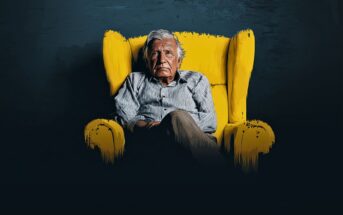 An elderly man with gray hair sits pensively in a bright yellow armchair against a dark background, wearing a striped shirt and khaki pants, with his arms folded and legs crossed.
