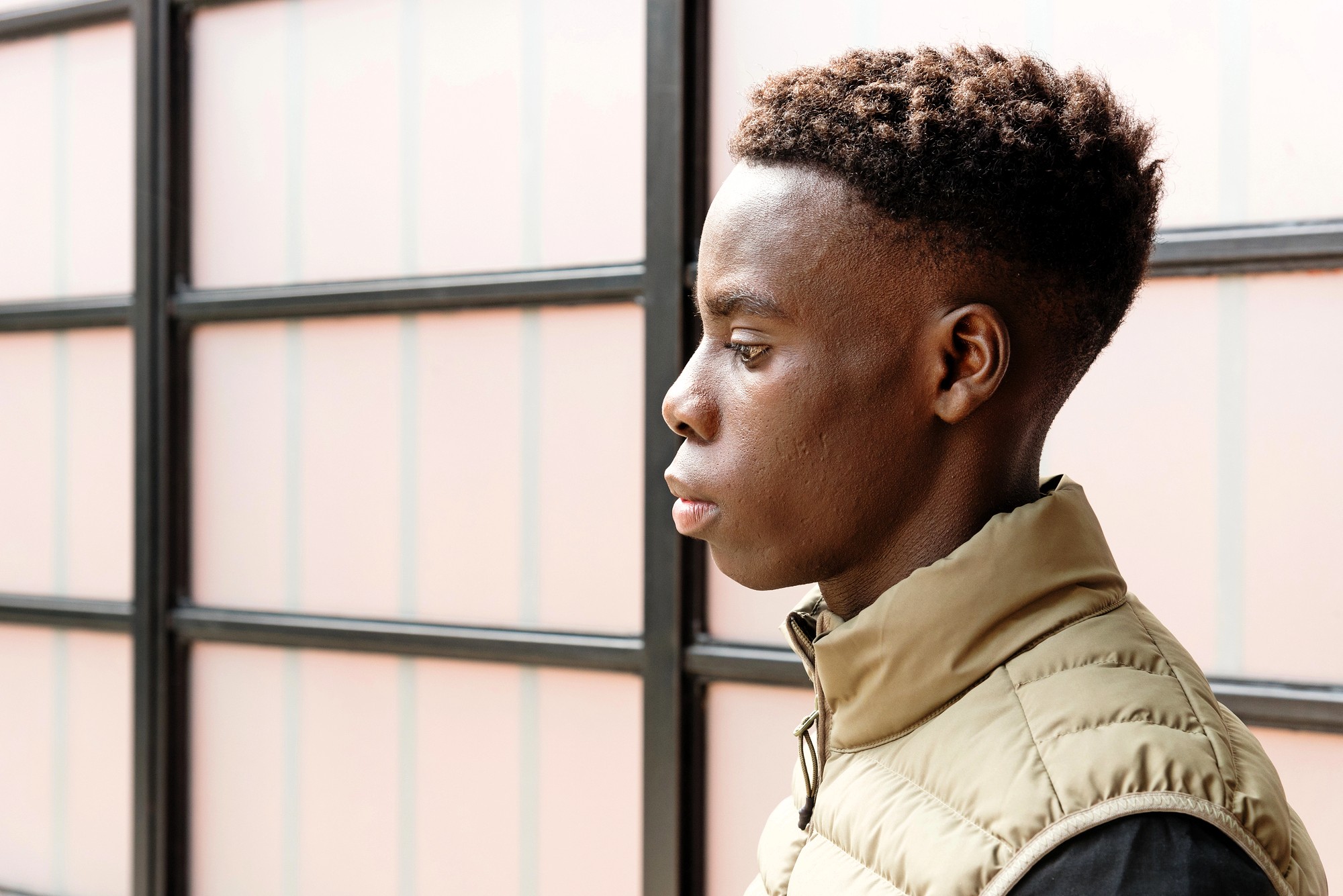 A young person with short, curly hair is shown in profile, wearing a light tan vest over a dark shirt, standing in front of a window with frosted glass panels.