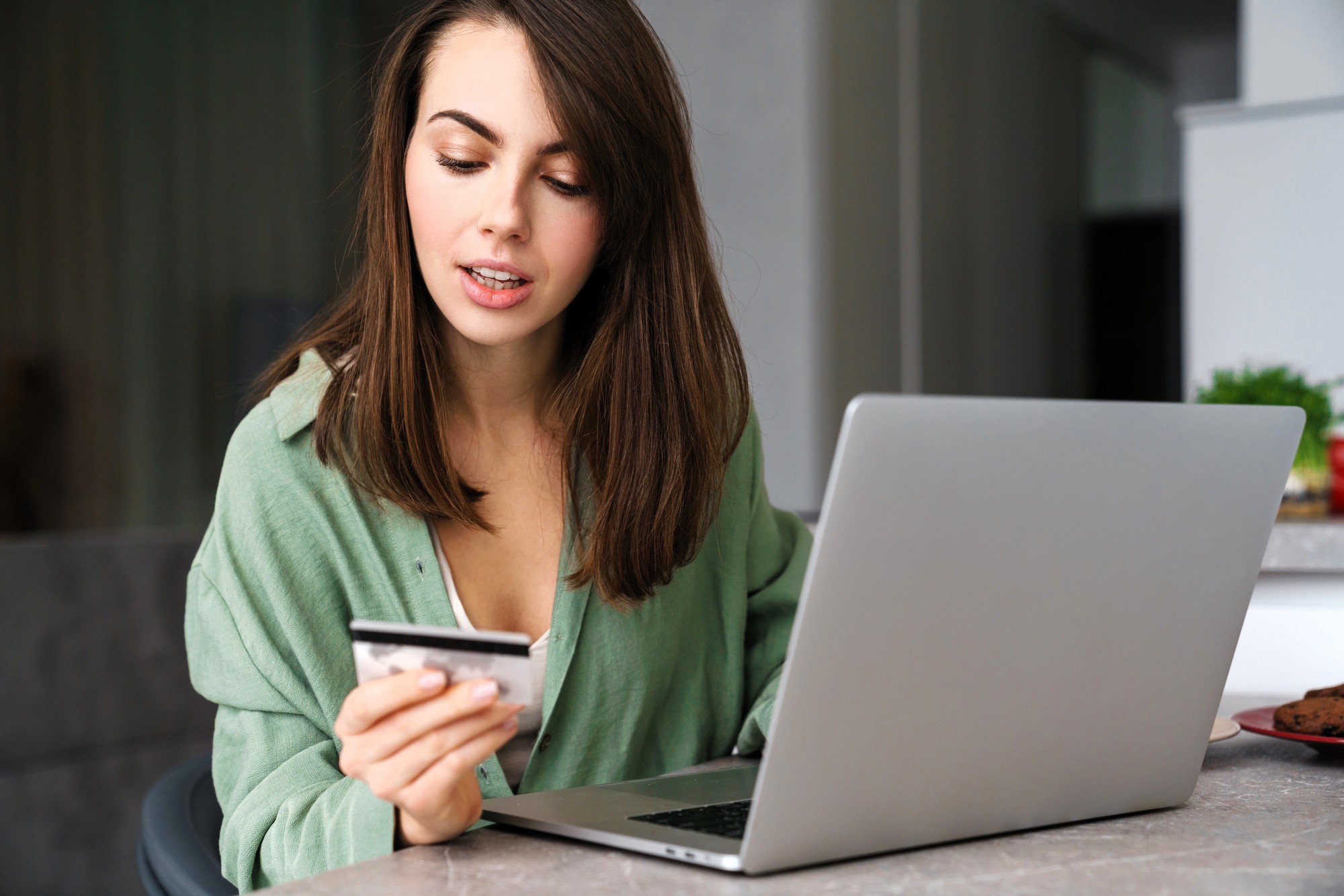 A woman with long brown hair sits at a table using a laptop and holding a credit card, appearing focused as she looks at the card. She is wearing a green shirt in a modern indoor setting.