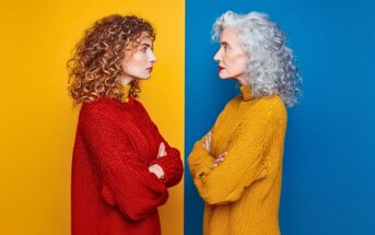 A young woman in a red sweater with curly hair faces an older woman in a yellow sweater with gray curly hair; they stand against contrasting yellow and blue backgrounds, both with arms crossed.