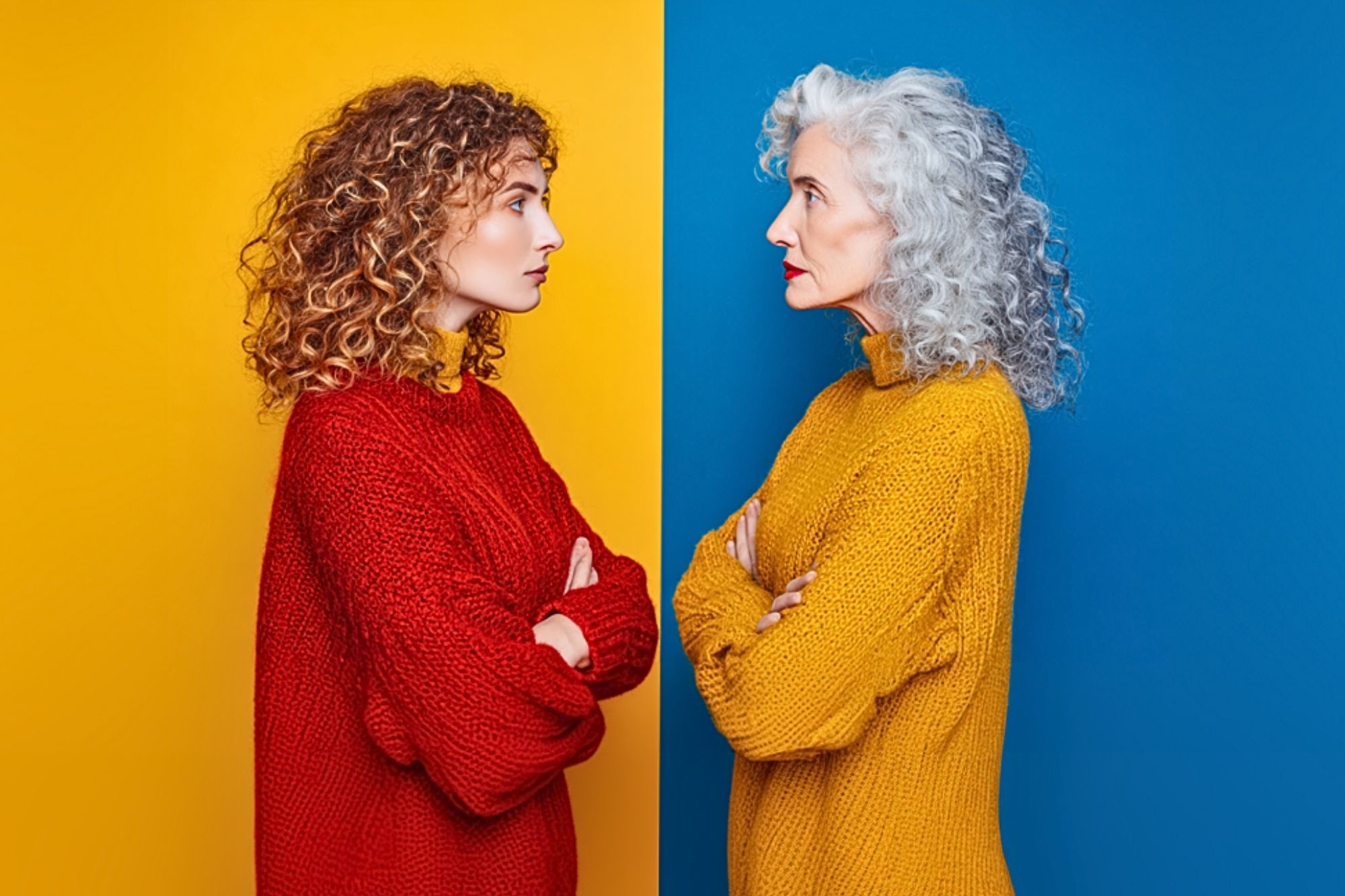 A young woman in a red sweater with curly hair faces an older woman in a yellow sweater with gray curly hair; they stand against contrasting yellow and blue backgrounds, both with arms crossed.