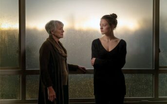 An older woman and a younger woman stand by a rain-covered window at sunset, facing each other. The older woman looks at the younger, who gazes outside with arms crossed, suggesting a serious or emotional moment.