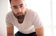 A man with a full beard and short hair wearing a light-colored henley shirt leans forward, looking directly at the camera. The background is bright and softly out of focus.