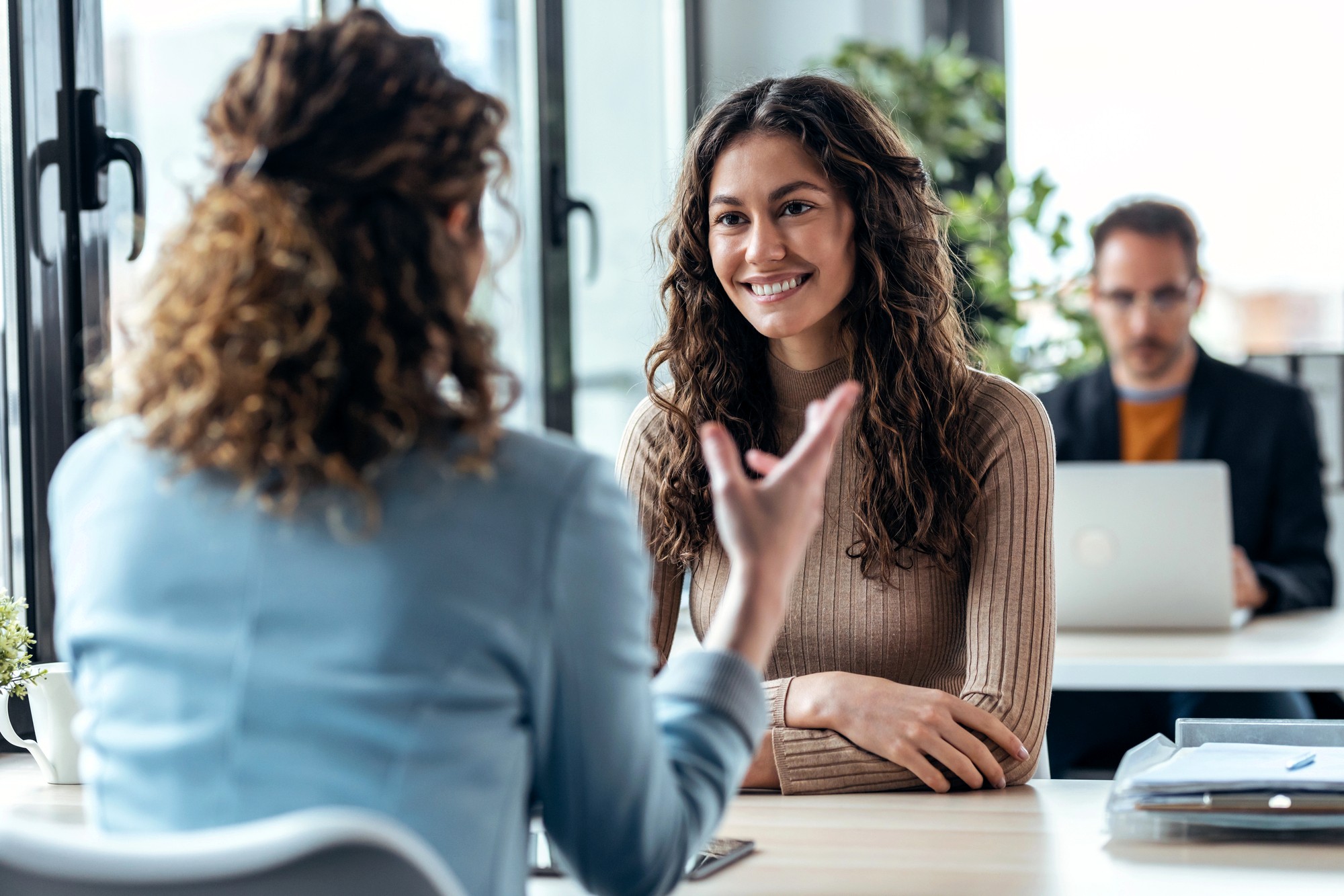 Two women sit across from each other at a desk, engaged in conversation. One woman smiles while listening, while a man works on a laptop in the background. The setting appears to be a modern office.