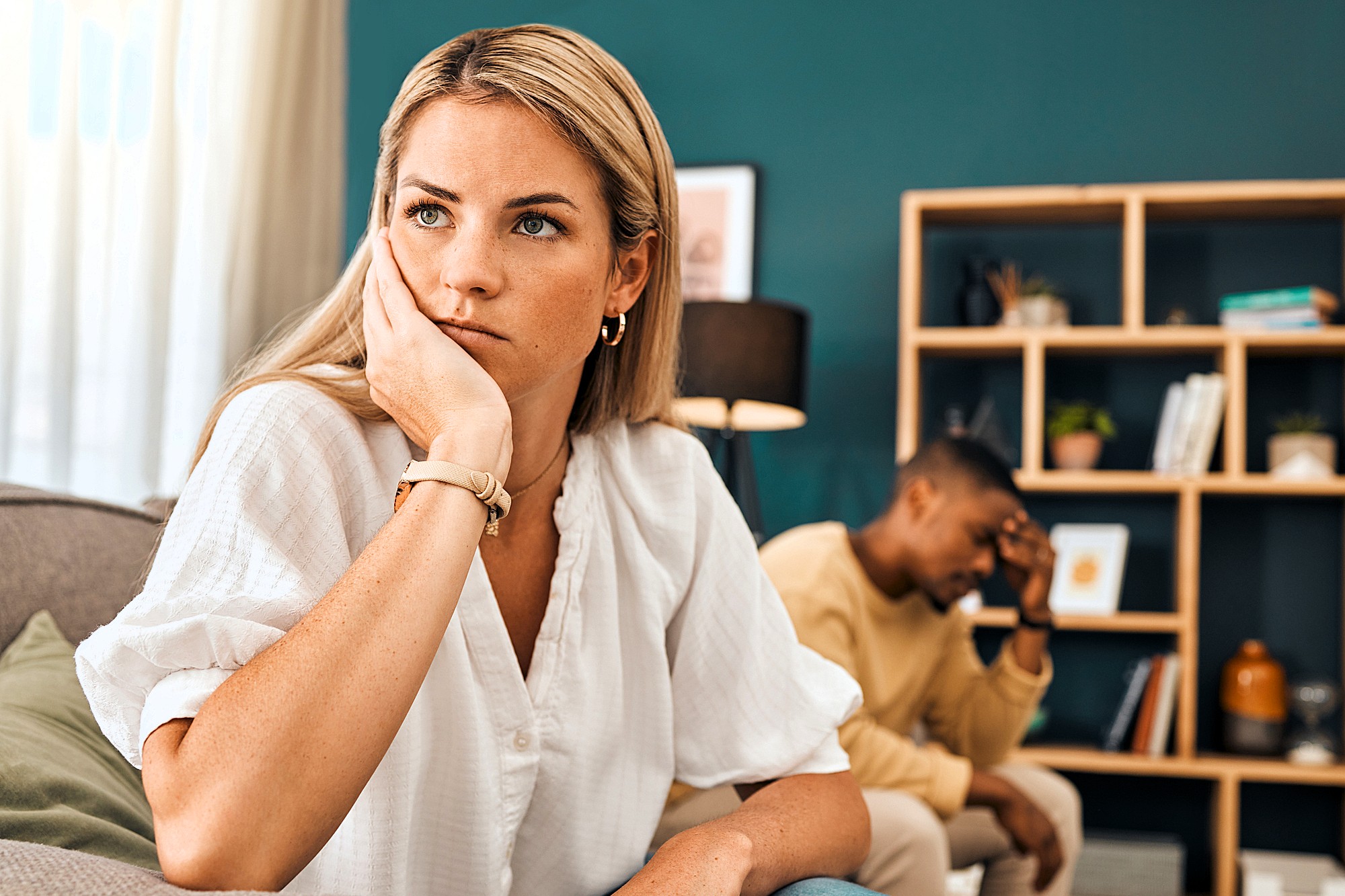 A woman sits on a couch looking away with a thoughtful, concerned expression, resting her face on her hand. In the background, a man sits with his head in his hand, appearing upset. Shelves and home decor are visible.