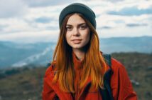 A young woman with long red hair, wearing a gray beanie and a red jacket, stands outdoors with a blurred landscape of hills and cloudy sky in the background.