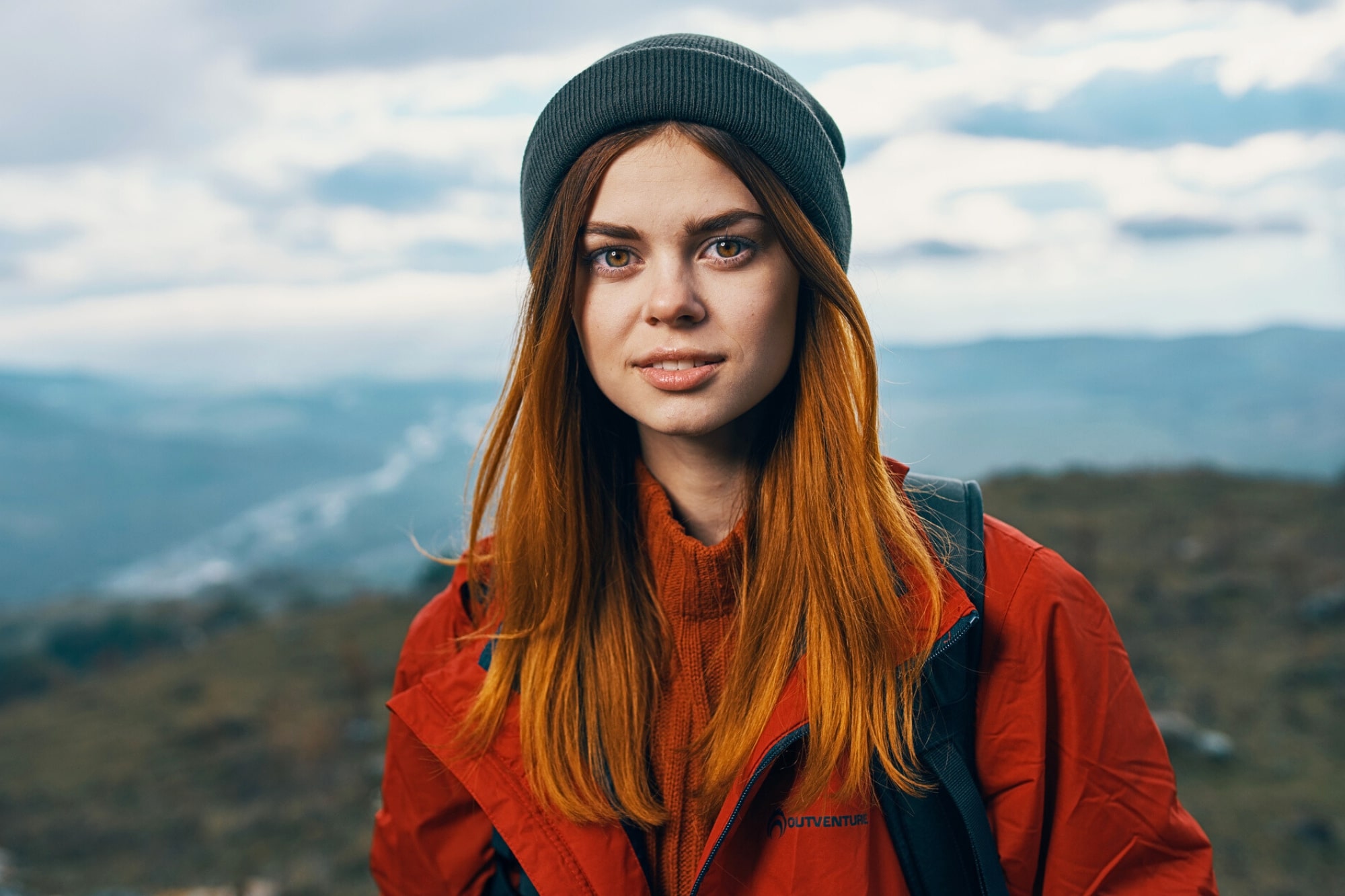 A young woman with long red hair, wearing a gray beanie and a red jacket, stands outdoors with a blurred landscape of hills and cloudy sky in the background.