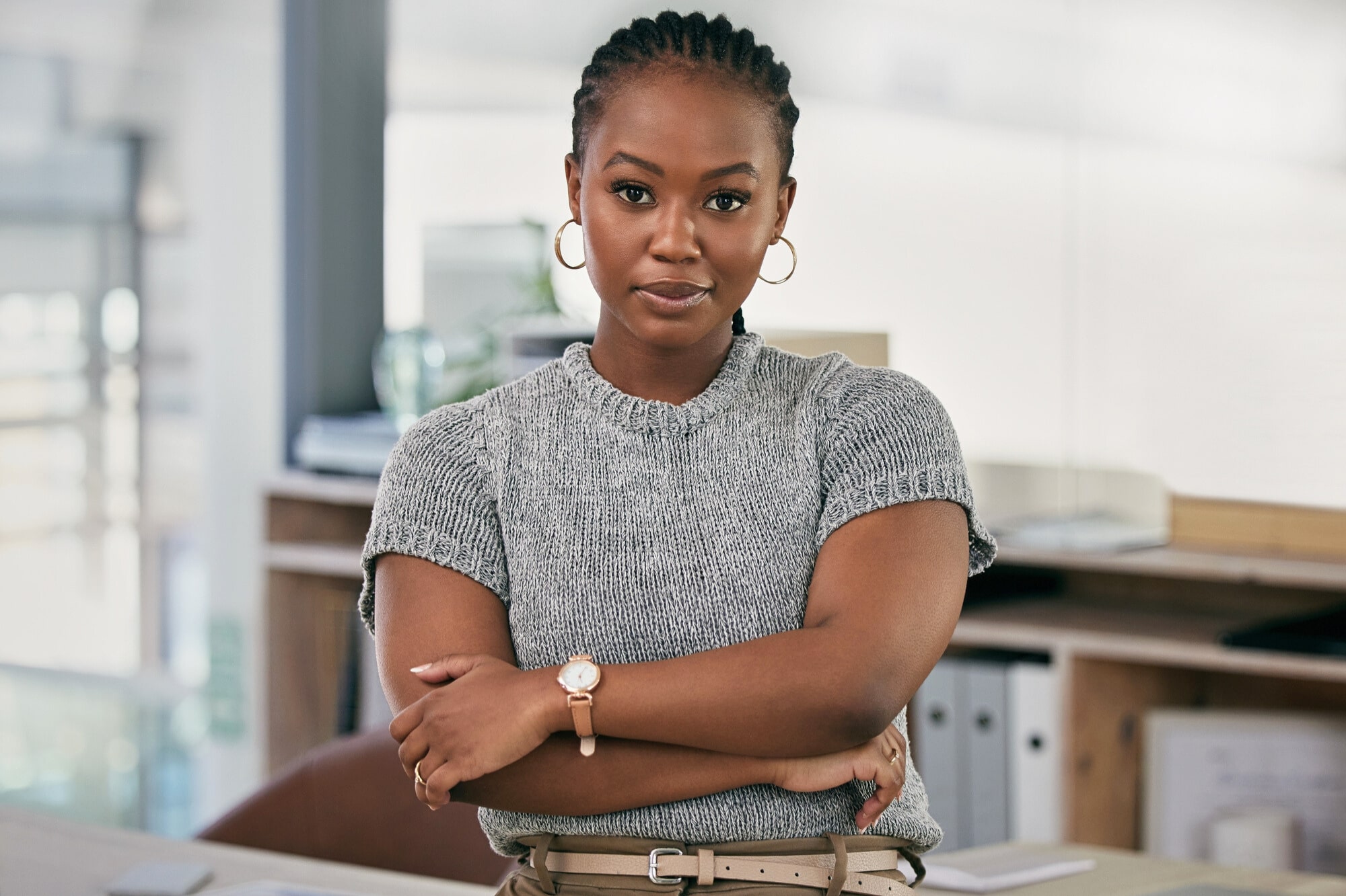 A woman with braided hair, wearing a grey short-sleeved sweater, hoop earrings, and a watch, stands with arms crossed in an office setting, looking confidently at the camera. Shelves and office supplies are in the background.