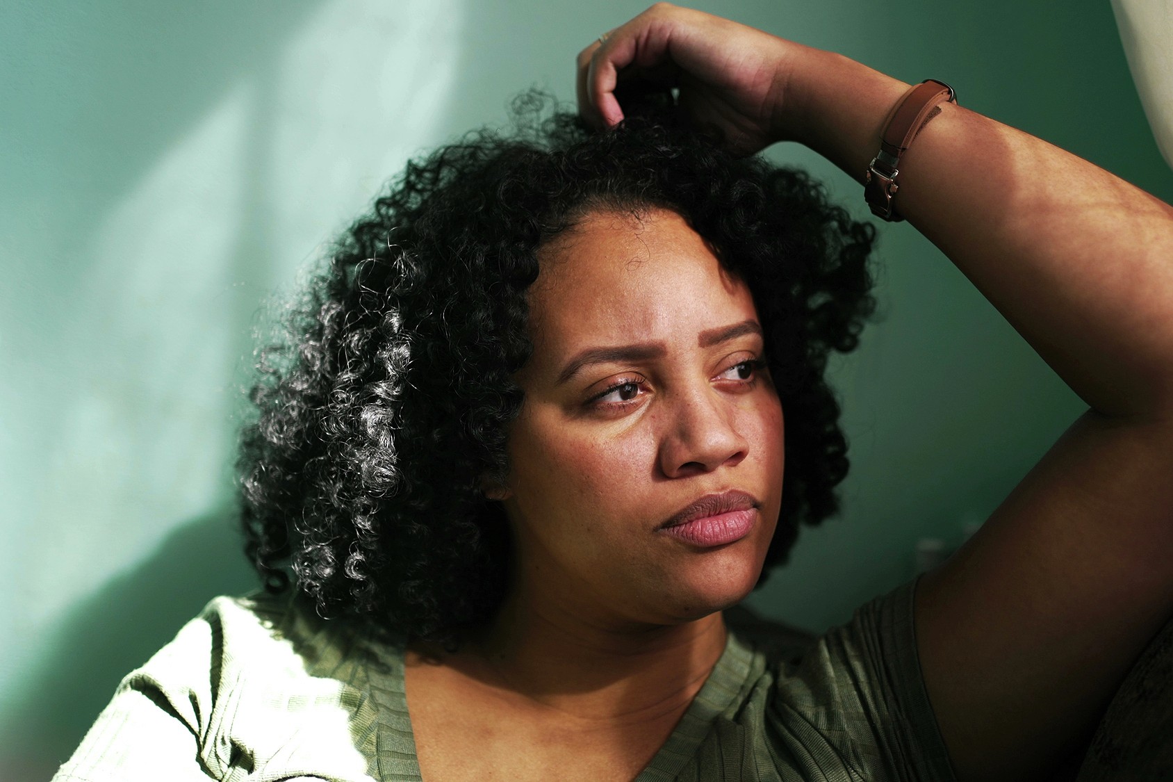 A woman with curly hair sits in soft sunlight, leaning against a green wall with her arm resting on her head, looking thoughtful and slightly concerned.