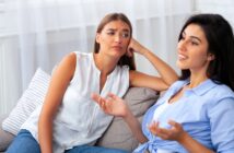 Two women sitting on a couch talking. One woman is speaking and gesturing with her hands, while the other looks bored or uninterested, resting her head on her hand. They are in a bright, cozy room.