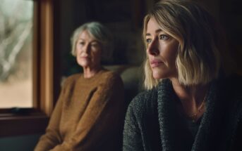 Two women sit indoors by a window. The woman in the foreground with blonde hair looks pensive, while an older woman with gray hair and a brown sweater sits in the background, slightly out of focus.